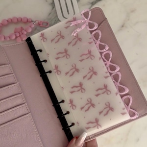 May include: A pink planner with a clear overlay patterned with pink bows. The planner has a pink bow embellishment and a pink beaded bracelet. The planner is open, showing card slots and a black spine.