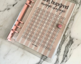 May include: A clear plastic binder with a pink ring mechanism holds a pink and white striped sheet with the text "one hundred envelope challenge" and a grid of numbers from 1 to 100.