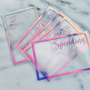 May include: A collection of clear budget envelopes with iridescent pink and purple frames. Each envelope is labelled with a different term in cursive script, such as "Gas", "Left Overs", "Groceries", and "Spending".