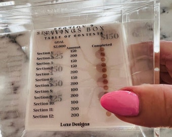 May include: A clear acrylic box with a chart inside that shows a savings goal of $2,000 and a completed amount of $150. The chart is divided into 12 sections, each with a dollar amount and a corresponding number of small brown circles to represent the amount saved. The chart is labeled "Section 2 Savings Box Table of Contents".