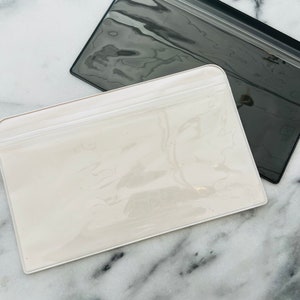 May include: Two clear plastic zippered pouches, one white and one black, on a marble surface.