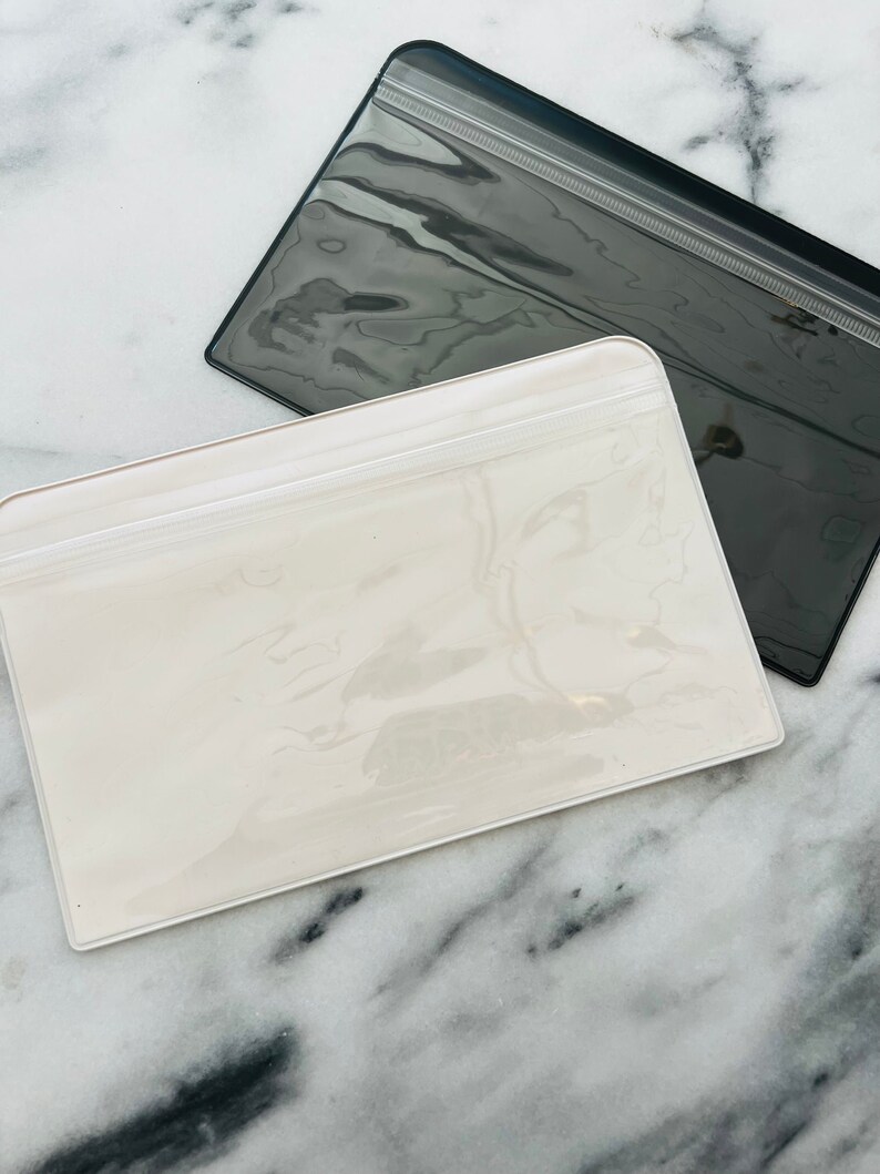 May include: Two clear plastic zippered pouches, one white and one black, on a marble surface.