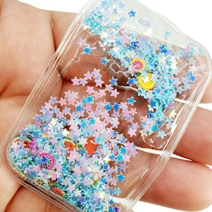 May include: A clear plastic pouch filled with blue and pink glitter stars and colorful fruit charms. The pouch is sealed and has a smooth, shiny surface.