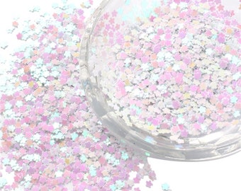 May include: A clear container filled with iridescent white and pink flower-shaped glitter. The glitter is scattered around the container.