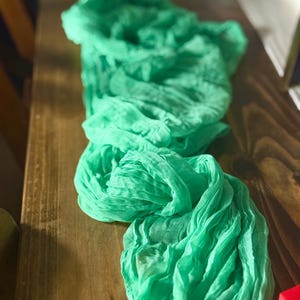 May include: A length of bright green fabric, possibly gauze or a similar lightweight material, is draped across a wooden surface. The fabric is bunched and gathered, creating a textured, flowing appearance. The colour is a vibrant, spring-like green.