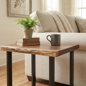 May include: A small, live-edge wooden side table with black metal legs. A potted fern, a stack of books, and a gray mug sit on the table. The table is next to a beige sofa.