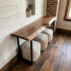 May include: A walnut wood console table with a live edge and a black metal frame. Three woven poufs are positioned beneath the table. The table is against a white shiplap wall and a brick wall. A decorative jug and dried lavender are in a niche.