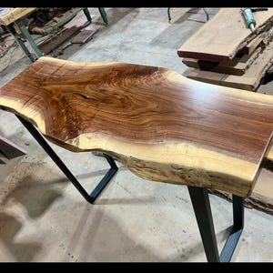 Bring the warmth of the Ozarks into your home with this stunning, handcrafted live-edge black walnut console table. Featuring an exceptionally unique, swirling grain pattern and the beautiful natural contrast between dark heartwood and creamy sapwood, this piece serves as a timeless functional art piece for any entryway, hallway, or behind-the-sofa display. Paired with sleek, modern industrial metal legs, it perfectly balances rustic charm with contemporary design.