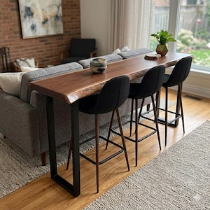 May include: A dark wood bar table with a live edge design, supported by black metal legs, is placed behind a grey sofa. Three black bar stools with wooden legs are positioned in front of the table. A small plant and a bowl are on the table.