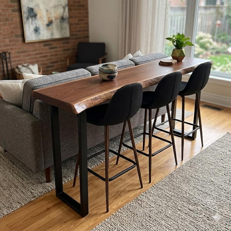 May include: A dark wood bar table with a live edge design, supported by black metal legs, is placed behind a grey sofa. Three black bar stools with wooden legs are positioned in front of the table. A small plant and a bowl are on the table.