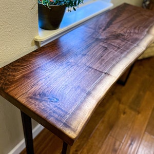 May include: A long, narrow, live edge walnut wood table with a dark brown finish. The table has a natural, organic shape and is supported by two black metal legs.