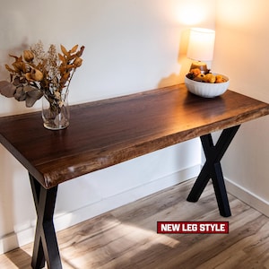 May include: A dark brown wooden table with a live edge top and black metal X-shaped legs. The table is decorated with a vase of dried flowers and a bowl of fruit. The text "NEW LEG STYLE" is visible.