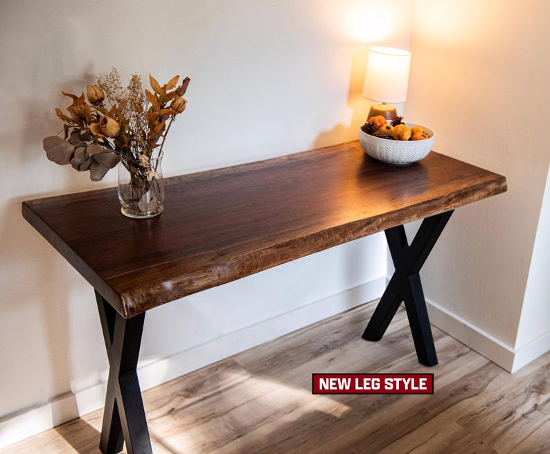 May include: A dark brown wooden table with a live edge top and black metal X-shaped legs. The table is decorated with a vase of dried flowers and a bowl of fruit. The text "NEW LEG STYLE" is visible.