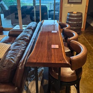 May include: A live-edge wooden bar with a dark finish, supported by black metal legs. Three brown leather bar stools are positioned along the bar. A brown wooden barrel is in the background. The bar is next to a brown leather sofa.