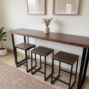 May include: A dark wood bar table with three matching stools. The table has a rectangular top and black metal legs. The stools have square seats and black metal frames. A small vase with dried flowers sits on the table.