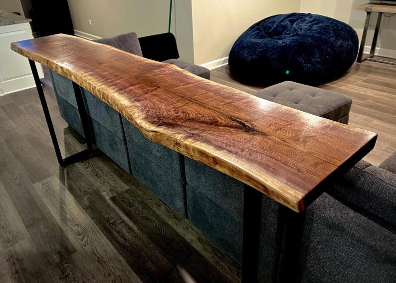 May include: A wooden sofa table with a live edge, featuring a warm brown hue. The table is supported by black metal legs and sits atop a grey sofa. The natural edge of the wood gives a unique, rustic feel to the piece.