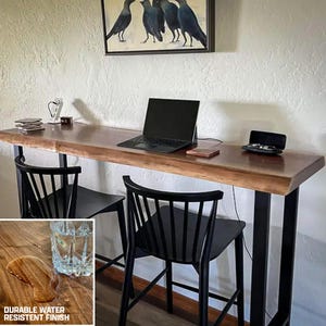 May include: A live edge wooden bar table with black metal legs, two black chairs, and a laptop. The table has a durable water-resistant finish. A painting of crows hangs above the table.