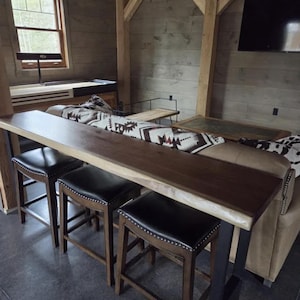 May include: A wooden bar with a live edge design and three black leather stools. The bar has a dark brown finish and is supported by metal legs. A patterned blanket is on a sofa nearby.