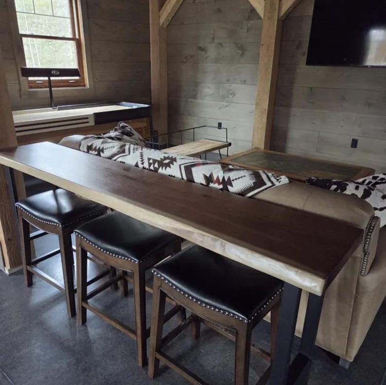 May include: A wooden bar with a live edge design and three black leather stools. The bar has a dark brown finish and is supported by metal legs. A patterned blanket is on a sofa nearby.