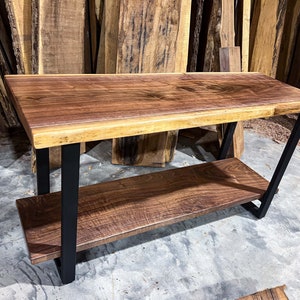 May include: A handcrafted wooden coffee table with a dark brown tabletop and a lower shelf. The table has black metal legs and a natural wood edge. The dimensions are approximately 48 inches long, 24 inches wide, and 18 inches high.