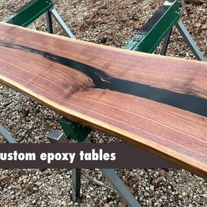 Handcrafted American black walnut live-edge table top featuring a smooth black epoxy resin river inlay. This premium, artisanal wood slab showcases deep chocolate grain patterns and natural live edges, perfect for a modern farmhouse console table or custom dining piece. Milled and finished in the Ozark Mountains