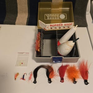 Puede incluir: Una caja de aparejos de pesca abierta con varios señuelos y flotadores. La caja es gris y beige, con la inscripción "BOBBER STOP". Varios señuelos coloridos con anzuelos están colocados frente a la caja, junto con dos flotadores blancos y rojos.