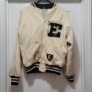 May include: Cream-colored varsity jacket with black and white striped trim. Features a large black and white letter "E" patch, a smaller "E" patch, and the number "71". The jacket has a button-up front and ribbed cuffs.