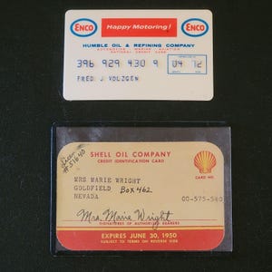 Puede incluir: Dos tarjetas de crédito antiguas. La tarjeta superior es de ENCO, con el texto "Happy Motoring!" y un número de tarjeta. La tarjeta inferior es una tarjeta de crédito de Shell Oil Company, con el nombre "Mrs. Marie Wright" y una fecha de caducidad.