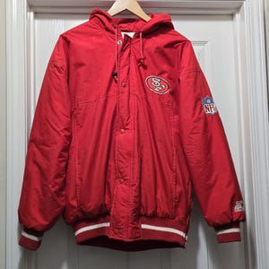 May include: Red hooded jacket with the San Francisco 49ers logo on the chest and NFL patch on the sleeve. The jacket has a white and red striped hem and cuffs. The jacket is made of a shiny material.