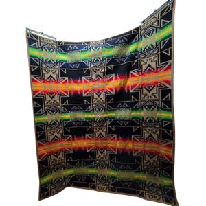 May include: A large, rectangular blanket with a geometric pattern in shades of black, tan, red, orange, yellow, and green. The blanket features a central design with symmetrical shapes and a border in a contrasting color.