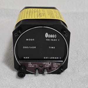 May include: A black Tri-Nav C navigation instrument with a circular display and various labeled controls. The display shows the text "00000" and other labels like "MODE", "OBS/VOR", "NAV", "TIME", and "EXT. LORAN C". A yellow label is affixed to the top.