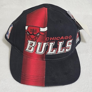 Puede incluir: Una gorra de béisbol negra con una franja roja vertical en el centro, con el logotipo y el texto de los Chicago Bulls bordados en blanco y rojo. La gorra incluye un parche con el logotipo de la NBA en el lateral y una etiqueta de papel.
