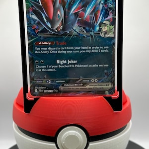 May include: A Pokémon trading card featuring N's Zoroark ex, displayed in a clear protective case. The card has a colorful illustration of a creature with red and blue accents. The card is held in a red and white Poké Ball display stand.