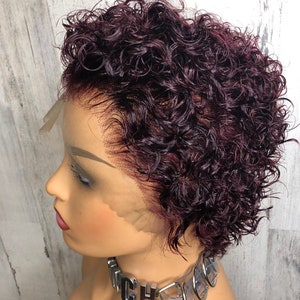 May include: A mannequin head with a short, curly, burgundy wig. The wig has a natural-looking hairline and is styled in a pixie cut. The mannequin is wearing a silver-toned choker necklace with the letters "CH" visible. The background is a light gray and white wood-paneled wall.