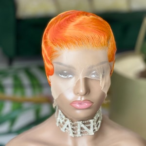 May include: A short orange wig with a side part and a wavy style. The wig is on a mannequin head with a sheer mesh cap. The mannequin is wearing a silver choker necklace with the word "QUEEN" spelled out in rhinestones.