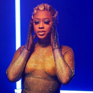 May include: A woman with blonde hair styled in finger waves wears a gold sparkly mesh top with long sleeves. She is posing with her hands on her face, and the background is a bright blue.