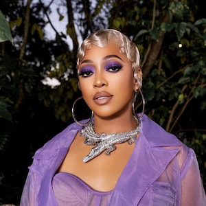 May include: A woman with short, platinum blonde hair styled in finger waves wears a purple sheer jacket and a silver alligator necklace. She has on purple eyeshadow and a nude lip.