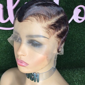 Peut inclure: Une perruque courte de couleur brun foncé avec une frange en dentelle et un style ondulé. La perruque est sur une tête de mannequin avec un collier ras du cou transparent qui dit "OUCH".
