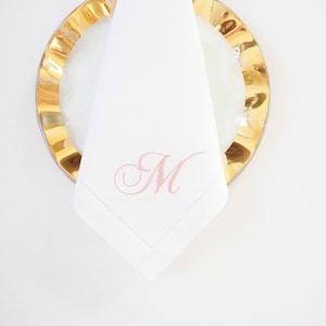 May include: A white linen napkin with a pink embroidered letter "M" on a gold plate.
