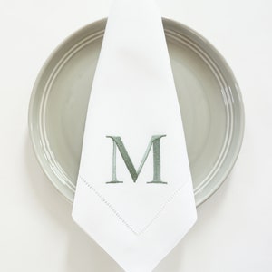 May include: White cloth napkin with a green embroidered letter "M" and a decorative stitched border, resting on a gray plate. The napkin is folded in a triangle shape, creating a formal table setting.