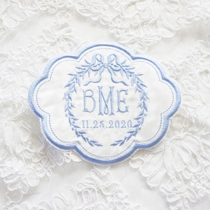 May include: A white embroidered patch with a blue border. The patch features a wreath of leaves, a bow, and the initials "BME" with the date "11.25.2020" below.