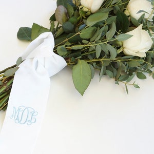 May include: A white bouquet of white roses and greenery tied with a white satin ribbon with the monogram "M.O.B." embroidered in blue.