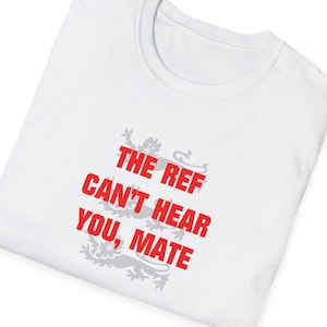 May include: White t-shirt with the text "THE REF CAN'T HEAR YOU, MATE" in red, with a gray emblem below. The shirt is folded, showing the collar and a portion of the front. The text is in a bold, sans-serif font.