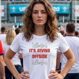 Puede incluir: Una camiseta blanca con el texto "IT'S GIVING OFFSIDE" en rojo, con un emblema gris. La camiseta la lleva una persona de pie frente a una multitud. El fondo incluye un estadio y banderas.