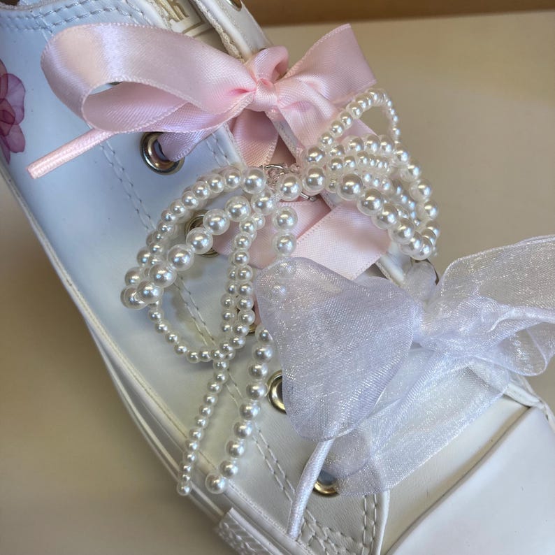 Puede incluir: Zapatillas blancas de lona con cordones de cinta de sat&eacute;n rosa atados en un lazo. Los cordones est&aacute;n decorados con un collar de perlas.
