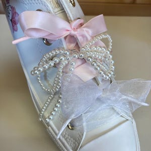 Puede incluir: Zapatillas blancas de lona con cordones de cinta de sat&eacute;n rosa, cuentas de perlas blancas y un lazo de tul blanco.