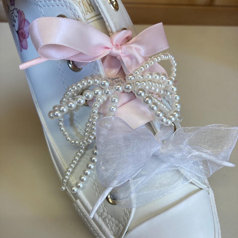 Puede incluir: Zapatillas blancas de lona con cordones de cinta de sat&eacute;n rosa, cuentas de perlas blancas y un lazo de tul blanco.