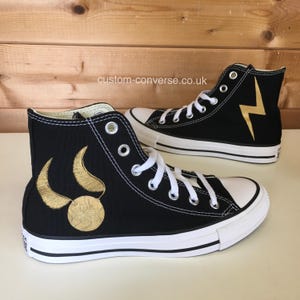 May include: A pair of black high-top Converse sneakers with custom designs. One shoe features a gold lightning bolt, while the other has a gold Snitch and wings from the Harry Potter series.