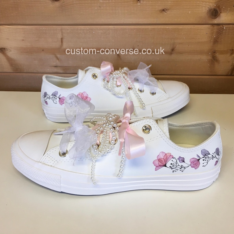 Puede incluir: Zapatillas blancas de lona con dise&ntilde;os florales rosas y adornos de perlas y cintas. Los zapatos est&aacute;n atados con una cinta rosa y tul blanco. El texto "custom-converse.co.uk" es visible en la imagen.