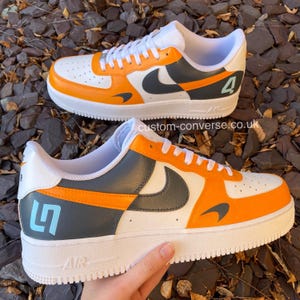 May include: Custom white, orange, and grey trainers with a white sole. The trainers have a grey Nike swoosh, orange detailing, and the number "4" in light blue on one shoe and "5" on the other. The website custom-converse.co.uk is visible.
